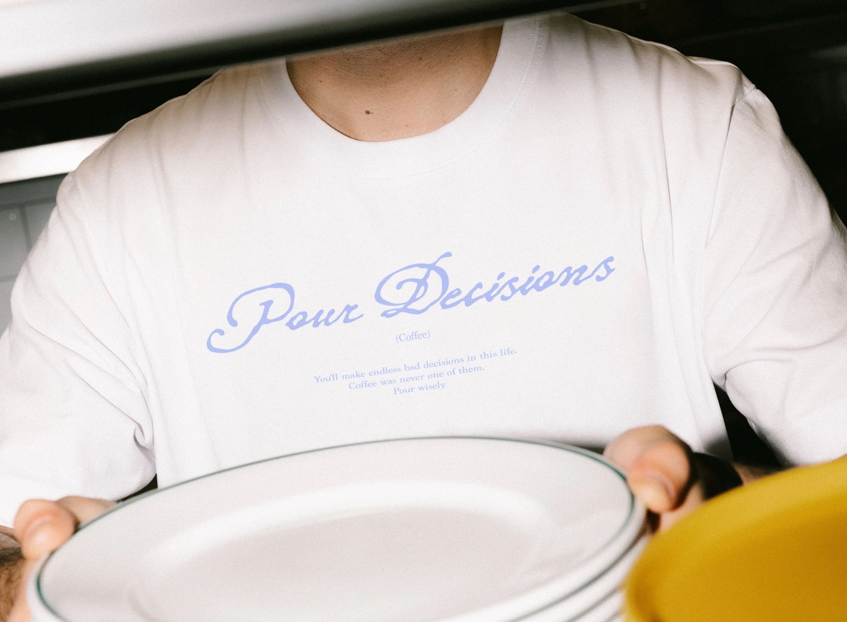 White t-shirt printed with the Pour Decisions script logo, worn by someone carrying plates